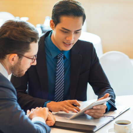 Closeup of two content adult business men sitting at table, using tablet computer, working with documents and discussing them in cafe